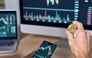 Man behind a computer with a phone in one hand and a bitcoin in the other.