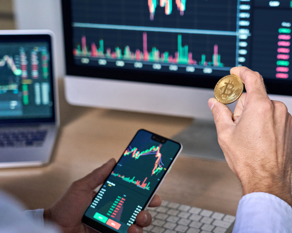 Man behind a computer with a phone in one hand and a bitcoin in the other.