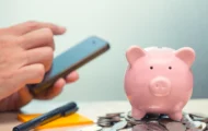 A piggy bank sitting atop of coins with a persons hand holding a phone to signify financial apps