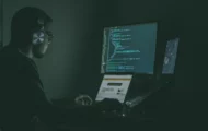 A man wearing headphones sits at a desk with two monitors, engrossed in his work.