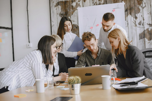 A diverse group of professionals collaborates around a laptop at a modern office table, with sticky notes and documents scattered around.