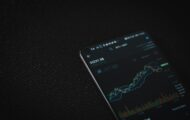 A smartphone displaying a cryptocurrency price chart and trading interface on a dark textured surface. The screen shows a rising candlestick chart with market indicators and trading volume, creating a dark, technology-focused financial atmosphere.